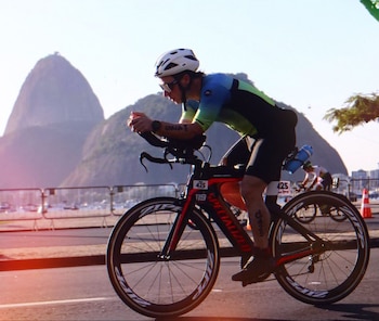 Un ciclista con casco blanco, gafas de sol y traje de colores pedalea una bicicleta negra y roja en una carretera. Al fondo, montañas y barcos