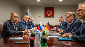 Hombres con traje sentados a ambos lados de una mesa de madera con pequeñas banderas de Rusia y Alemania, botellas de agua y cuadernos