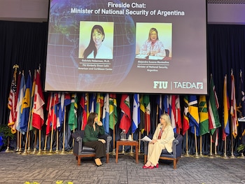 Dos mujeres sentadas en sillones frente a una pantalla con sus fotos e información, con un telón azul oscuro y banderas de países de fondo