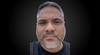 Hombre de tez morena, cabello oscuro entrecano, barba gris, bigote gris, y camiseta negra, posando de frente con un fondo oscuro.