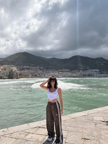 Griselda Siciliani parada em um penhasco com vista para o mar e uma cidade costeira com montanhas verdes sob um céu nublado. Use uma blusa branca e calça com estampa animal