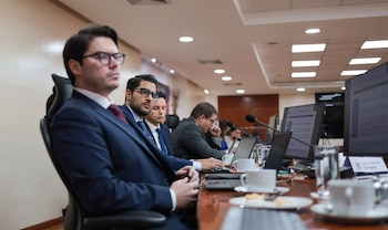 Hombres de traje sentados en una mesa. Hay laptops, monitores, micrófonos, tazas y vasos. Un cartel visible dice 'Javier Andres Alfaro Fitch Ratings'