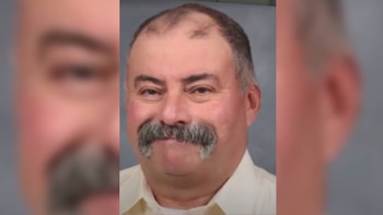 Primer plano de un hombre de mediana edad, tez clara, cabello oscuro escaso, cejas pobladas y bigote grisáceo grueso, con una sonrisa sutil