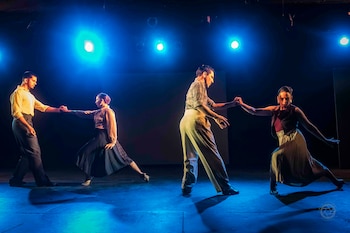 Cuatro bailarines en un escenario oscuro con luces azules intensas. Dos parejas se toman de las manos en poses dinámicas, vestidas con camisas y pantalones/faldas