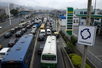 Tráfico denso de autobuses y automóviles en una avenida de Lima, con una gasolinera y un letrero de OSINERGMIN en primer plano.