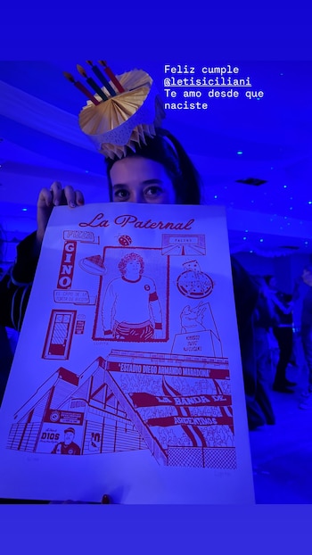 Persona con sombrero de cumpleaños y velas sostiene un póster rojo sobre fondo azul. El póster muestra ilustraciones de Diego Maradona y el estadio La Paternal