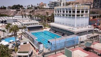 Vista aérea de complejo con piscina olímpica, piscina recreativa, canchas deportivas rojas, edificio moderno y palmeras bajo cielo claro
