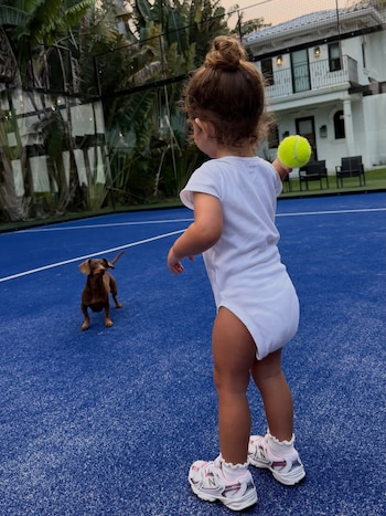 Niña de espaldas con moño y ropa blanca sostiene una pelota de tenis amarilla. Un perro salchicha marrón la mira en una cancha de pádel azul. Fondo con casa y plantas