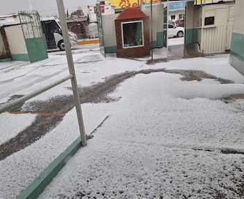 Calles y estacionamiento cubiertos por una densa capa de granizo blanco, con zonas húmedas y charcos de agua. Se ven edificios y un vehículo al fondo
