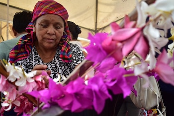 Una mujer mayor con un pañuelo de cuadros rojo, verde y azul mira hacia abajo, con el primer plano lleno de flores artificiales moradas, rosas y blancas