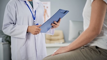 Doctora de bata blanca con acreditación azul sostiene un portapapeles azul, frente a paciente sentada de camiseta blanca y pantalón. Fondo difuso de consultorio.