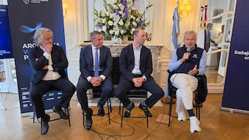 Cuatro hombres con vestimenta formal están sentados en sillas altas durante un evento junto a banderas nacionales