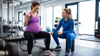 Una mujer embarazada en ropa deportiva morada levanta una mancuerna, sentada en un banco de gimnasio, junto a una entrenadora con chándal azul.
