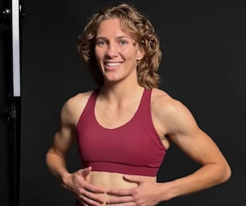 Una mujer joven de cabello rizado y tez clara sonríe, viste un top deportivo color rojo vino y tiene las manos sobre su abdomen. El fondo es oscuro