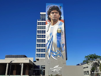 Mural vertical del rostro de Diego Maradona con camiseta celeste y blanca en la fachada de un edificio gris; cielo azul despejado y vegetación