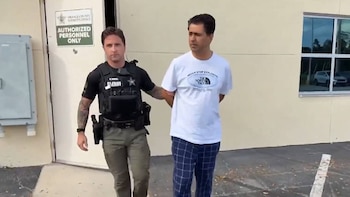 Un oficial del Sheriff del Condado de Orange escolta a un hombre con camiseta blanca y pantalones a cuadros, con manos detrás, fuera de un edificio policial