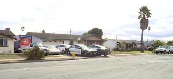 Dos vehículos policiales del Sheriff, varios automóviles, un camión de mudanzas, casas residenciales, un hidrante amarillo, palmeras y vegetación en una calle