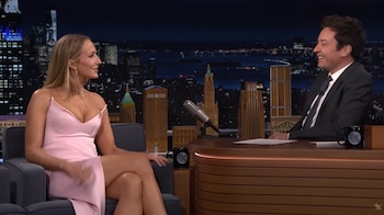 Michelle Yeoh, vestida con un elegante vestido rosa, está sentada en un sillón gris charlando y riendo con Jimmy Fallon en un estudio de televisión con un fondo urbano nocturno