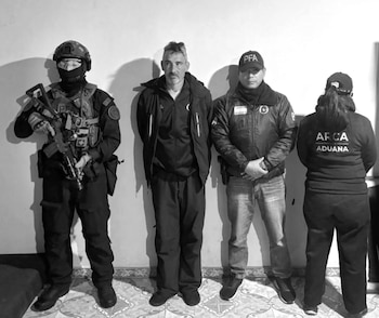 Un hombre de ropa oscura permanece de pie entre un agente táctico armado, un oficial con gorra de policía y una agente con chaqueta de Aduana