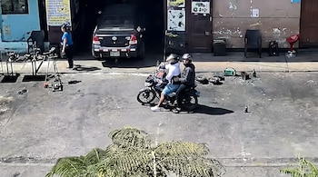 Dos personas con casco en una motocicleta sobre la calle frente a un taller mecánico con un automóvil negro y herramientas.
