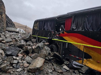 Vista lateral de un bus negro con franjas rojas, naranjas y amarillas rodeado por rocas de un deslizamiento. Un camión blanco se observa al fondo