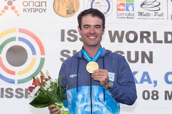Federico Gil sonríe mientras muestra una medalla dorada y sostiene un ramo de flores ante un panel promocional de la ISSF