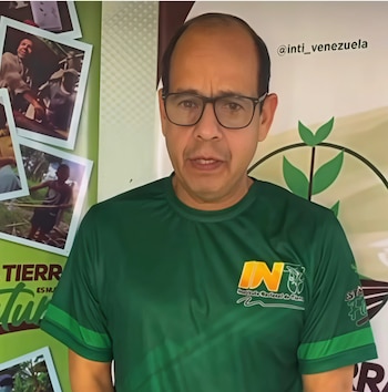 Hombre con anteojos y camiseta verde frente a un panel decorado con fotografías y una cuenta de red social