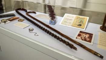Exhibición con dos bastones de madera, dos relojes de bolsillo, un tintero con pluma, un pasaporte abierto y fotografías sobre una superficie blanca