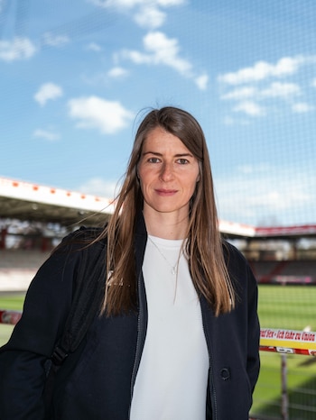 Mujer de cabello castaño y largo con chaqueta oscura y camiseta blanca, de pie frente a un estadio con cielo azul claro y nubes blancas