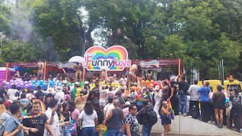 Multitud de personas en calle; carro alegórico con cartel luminoso de corazón arcoíris y letrero 'Funny Love'; hombres en tanga en la plataforma; globos; árboles verdes