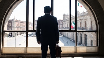 Un hombre de traje de espaldas sostiene un maletín frente a una ventana arqueada, con vista a una calle y el Palacio de Justicia con bandera mexicana.