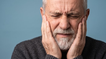 Primer plano de un hombre de edad avanzada con barba que sostiene sus mejillas con las manos y mantiene los ojos cerrados.