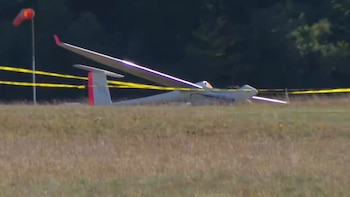 Un planeador blanco yace sobre la hierba en un área delimitada con cinta amarilla de seguridad, con árboles al fondo