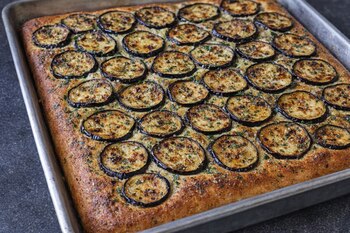 Focaccia de berenjena recién horneada y rectangular en una bandeja de metal. La superficie dorada está cubierta con rodajas de berenjena y hierbas.