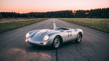 Un coche Porsche 550 Spyder plateado con el número 77 en el capó y laterales, con luces encendidas, en una pista de asfalto al atardecer.