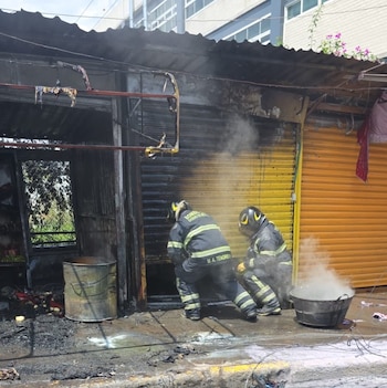 Dos bomberos con uniforme oscuro y casco están agachados frente a un puesto quemado, con humo saliendo. Hay un barril, una tina con humo y escombros