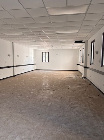 Interior de una sala vacía con paredes blancas, un techo de paneles suspendidos, ventanas rectangulares y un suelo de baldosas cubierto de tierra y escombros