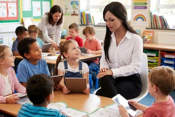 Robot humanoide de cabello oscuro y camisa blanca con niños en un aula escolar, algunos con tabletas, y una maestra al fondo