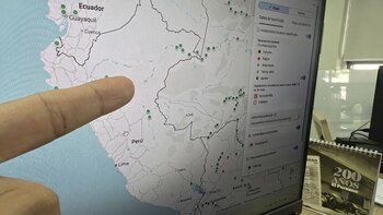Dedo de una mano apunta a un monitor que muestra un mapa de Sudamérica con marcadores verdes en Perú y Ecuador. Se ve un menú lateral y un calendario de mesa