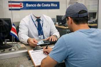 Un hombre con gorra escribe en un formulario de crédito. Otro hombre de camisa blanca sostiene una tarjeta de identificación. Hay una bandera de Costa Rica, un teclado y dinero.