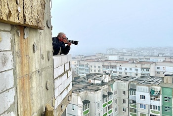 Hombre con gafas sosteniendo una cámara sobre el muro de un edificio, con bloques de apartamentos al fondo