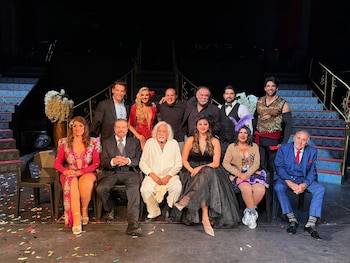 Doce actores con vestuarios de gala y trajes temáticos posan sentados y de pie frente a una escalera en el escenario de un teatro
