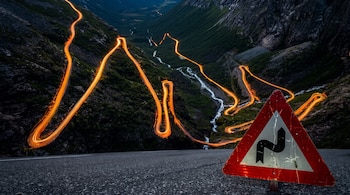 Señal de tránsito triangular roja con símbolo de doble curva. Carretera de asfalto en primer plano. Fondo con ruta de montaña, luces de vehículos y valle.