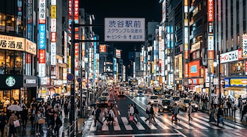 Calle urbana de noche con personas en un paso peatonal, taxis, letreros luminosos en idioma japonés y edificios a los lados.