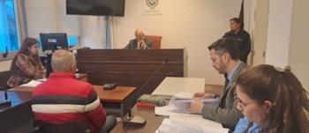 Una sala de audiencias judicial con un juez, tres personas sentadas en las mesas, una persona de pie y monitores de computadora