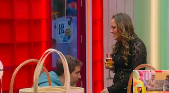 Un hombre con sudadera turquesa mira a una mujer con vestido negro de encaje que sostiene una copa en un set de televisión
