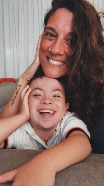 Mujer de cabello oscuro con piercings y una niña, ambas con grandes sonrisas, posan de cerca. La niña usa una camiseta polo blanca con rayas