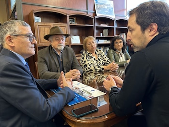Cinco personas sentadas alrededor de una mesa de madera. Se observan papeles, un bolígrafo y un teléfono. Hay estanterías de fondo con libros y objetos