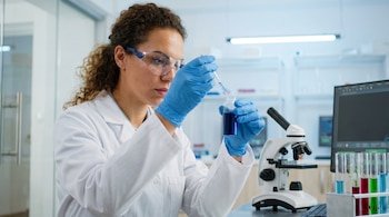 Una científica de pelo rizado y gafas de seguridad, con bata blanca y guantes azules, transfiere un líquido azul con un gotero en un laboratorio.