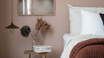 Un dormitorio con pared color malva, cama con ropa de cama blanca y marrón, taburete de madera con libros y un jarrón con flores secas, y lámpara colgante.